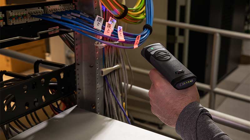 A worker at a server rack scans a qr code on a wrap around cable label using a barcode scanner.