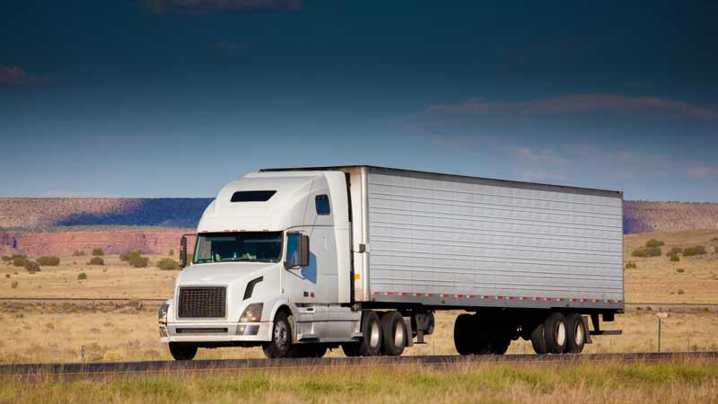 A profile shot of the side of a semi-truck that has DOT-compliant red and white reflective tape while driving down the highway.