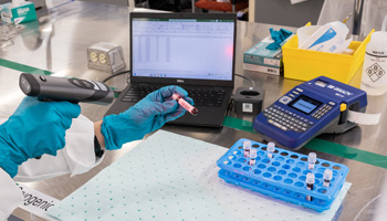 A person in a lab coat stands beside lab automation software, surrounded by labeled equipment and research materials.