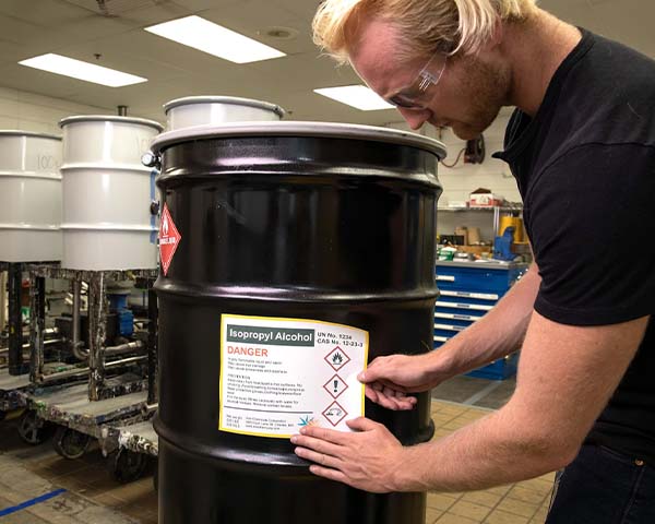 A man attaching an isopropyl alcohol GHS HazCom label to a barrel in a warehouse environment.